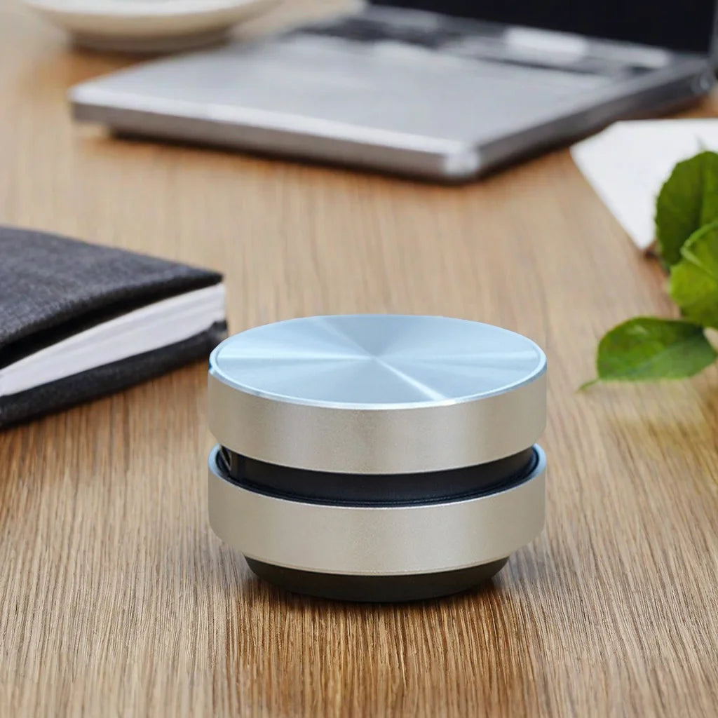 A compact and sleek SoundSurface Mini Resonance Speaker made of aluminum, displayed on a wooden surface, with a plant and a notebook in the background.